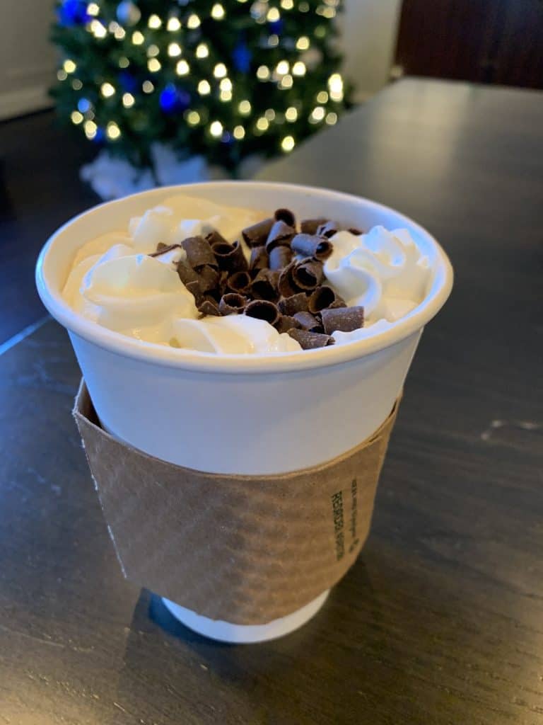 Cup of hot chocolate with whipped cream and chocolate shavings, with a Christmas tree in the background