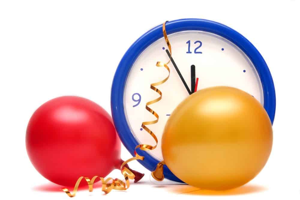 A red balloon and a gold colored balloon in front of a clock about to strike 12