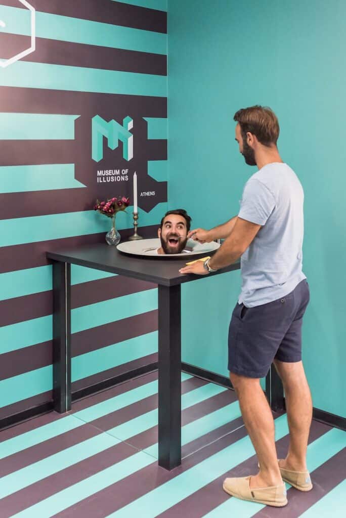 Man in green and brown striped room appearing to look at his own head on a plate