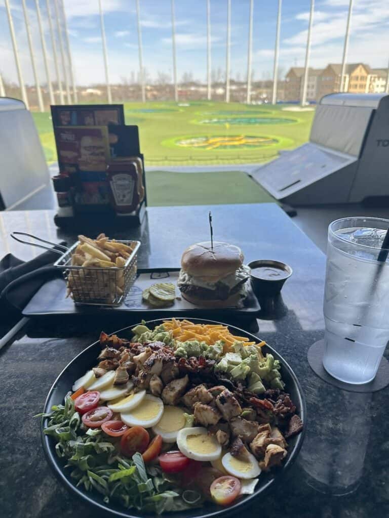 A plate of food and a drink on a table in front of a Topgolf range