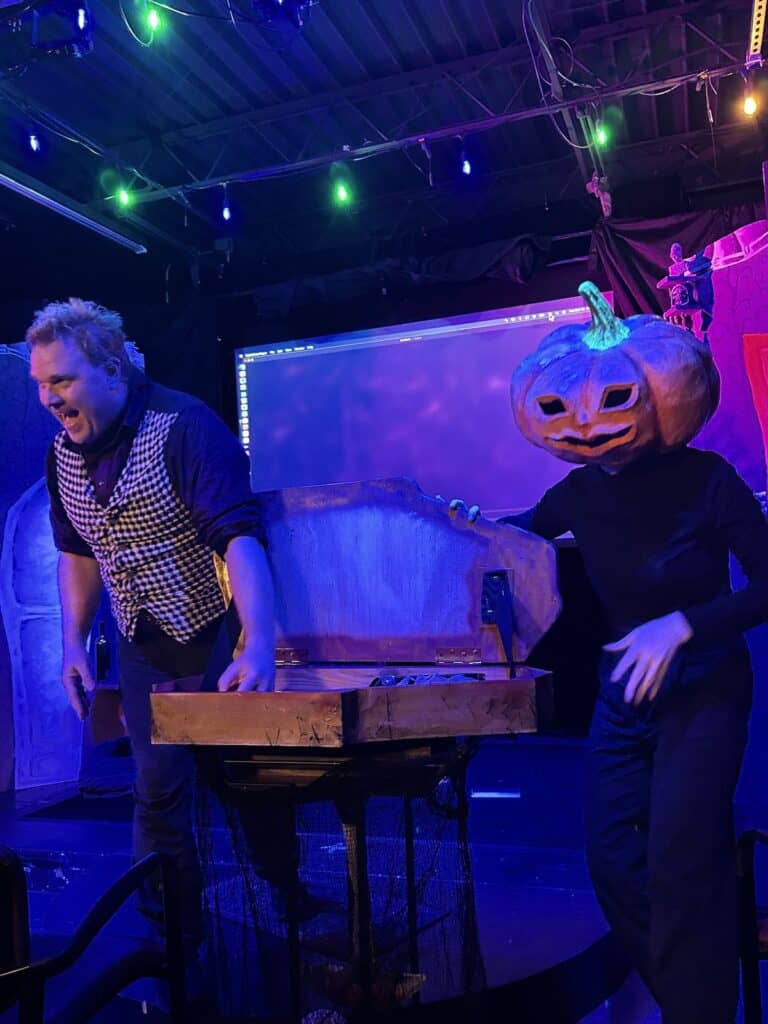 person with pumpkin head and person without pumpkin head standing with coffin of candy